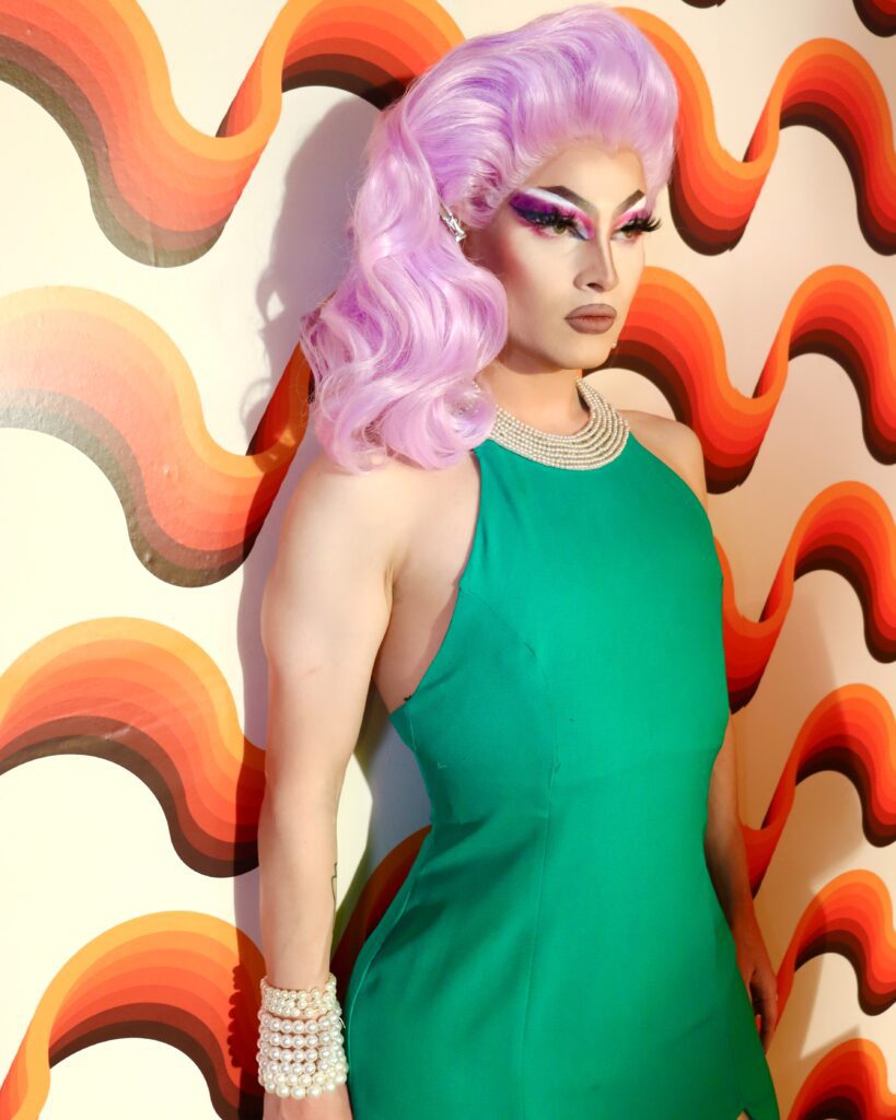 A woman in a green dress with dramatic makeup and a lavender wig stands in front of a white wall with orange wavy stripes