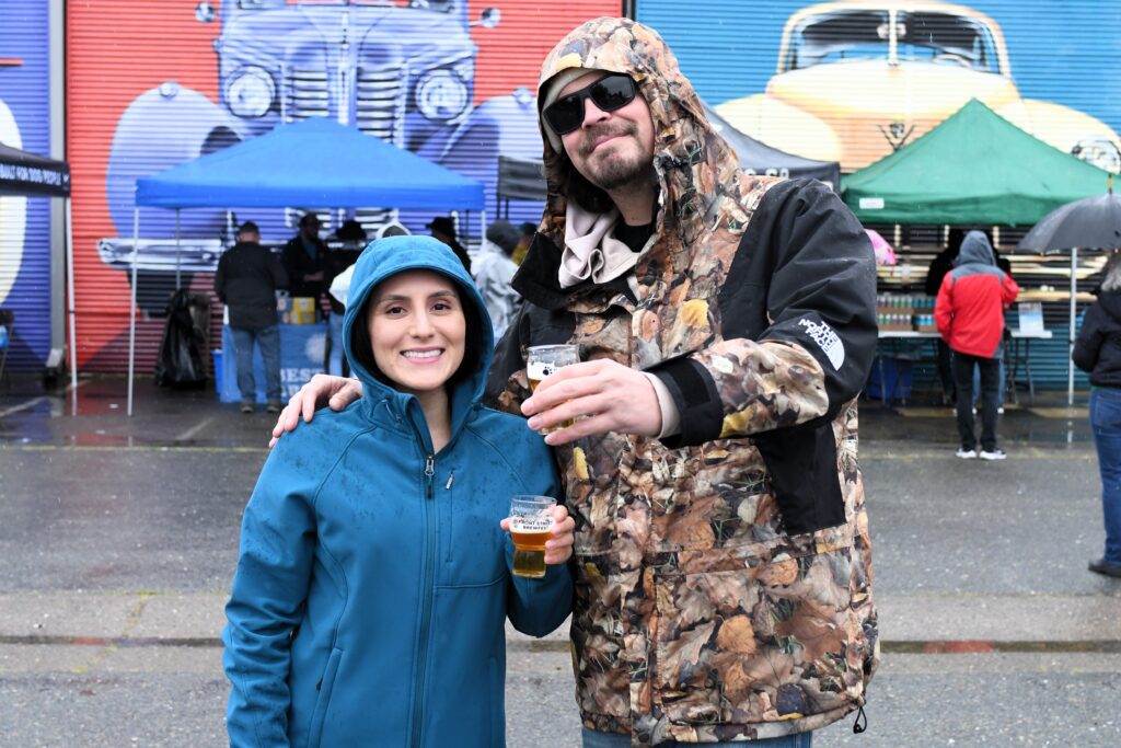 A tall person in camoflage jacket stands next to a shorter person in a blue rain coat. Both hold their beers toward the camera and smile