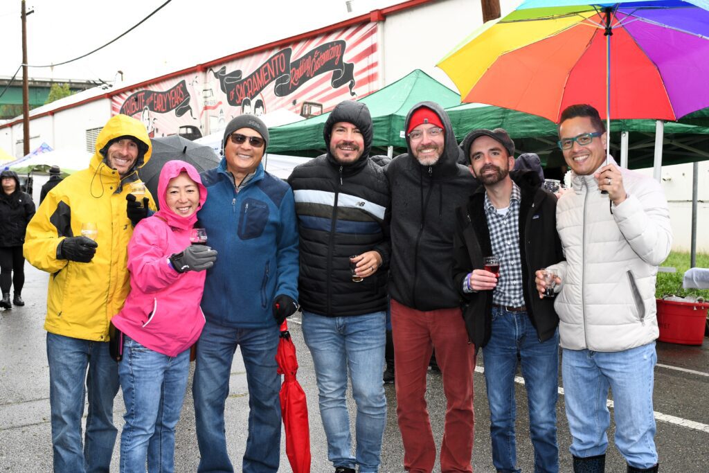 A group of seven people stand underneath a rainbow umbrella outside the California Auto Museum and raise their glasses