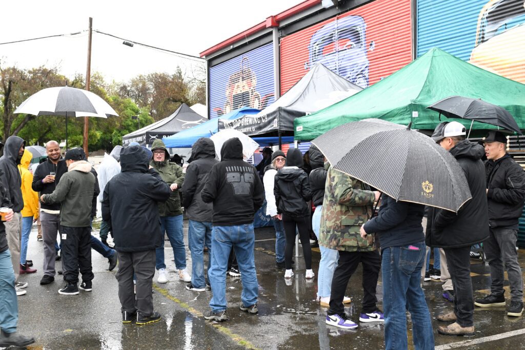 A crowd of people with umbrellas and rain gear visit tents outside the California Auto Museum