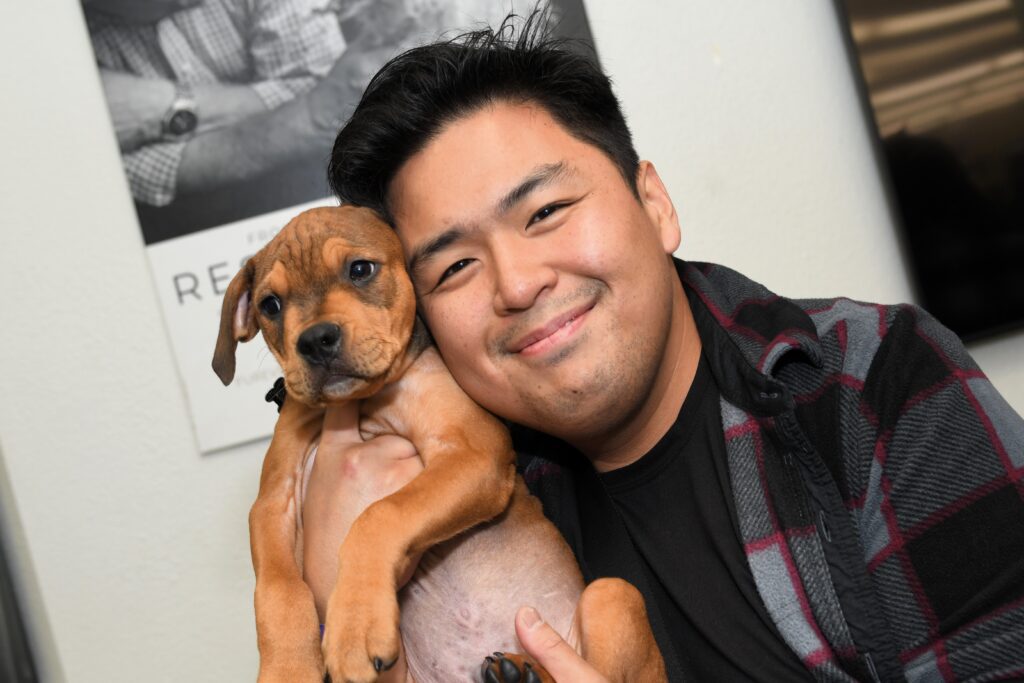 A smiling man holds a brown puppy up to his face